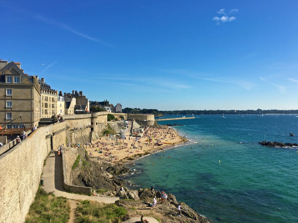 A coastal town features historic stone buildings and fortification walls along a curved sandy beach populated with beachgoers. The turquoise and blue waters meet rocky outcrops and golden sand, with swimmers visible in the shallow areas. Clear blue skies stretch across the scene, with the coastline extending into the distance where additional landmass and sailboats are visible on the horizon.
