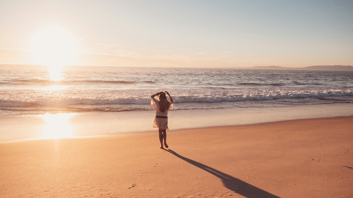 A person stands alone on a sandy beach facing the ocean, with their hands raised to their head, casting a long shadow on the sand. The scene is bathed in warm golden light from the sun positioned low on the horizon, with gentle waves rolling onto the shore and a distant landmass visible across the water. The lighting and color palette suggest either sunrise or sunset, creating a peaceful coastal atmosphere.
