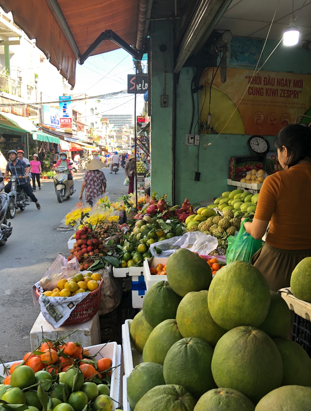 A bustling street market features a fruit vendor's stall in the foreground displaying a variety of tropical produce, including a prominent pyramid of large green pomelos, along with oranges, bananas, dragon fruit, and other fruits arranged in baskets and crates. Behind the stall, a busy street scene shows pedestrians, motorcyclists, and shopfronts with awnings lining both sides of the road. Vietnamese signage is visible on buildings, including advertising for products.
