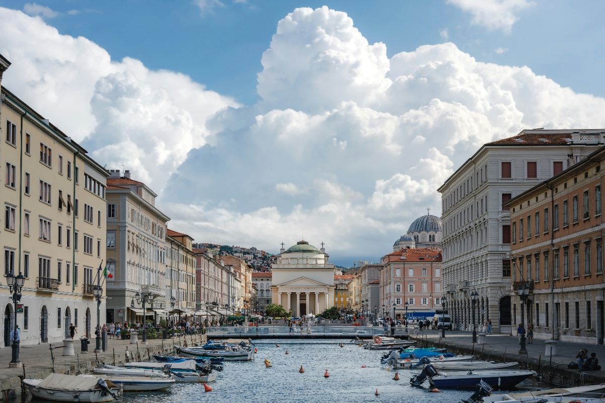 A canal lined with small boats runs through the center of a European coastal town, flanked by multi-story classical buildings in pastel colors. In the background, a neoclassical church or temple with columned portico and dome is centered at the end of the waterway, with hillside buildings visible beyond. Dramatic white cumulus clouds dominate a blue sky above the scene, while pedestrians occupy the waterfront plaza areas on both sides of the canal.