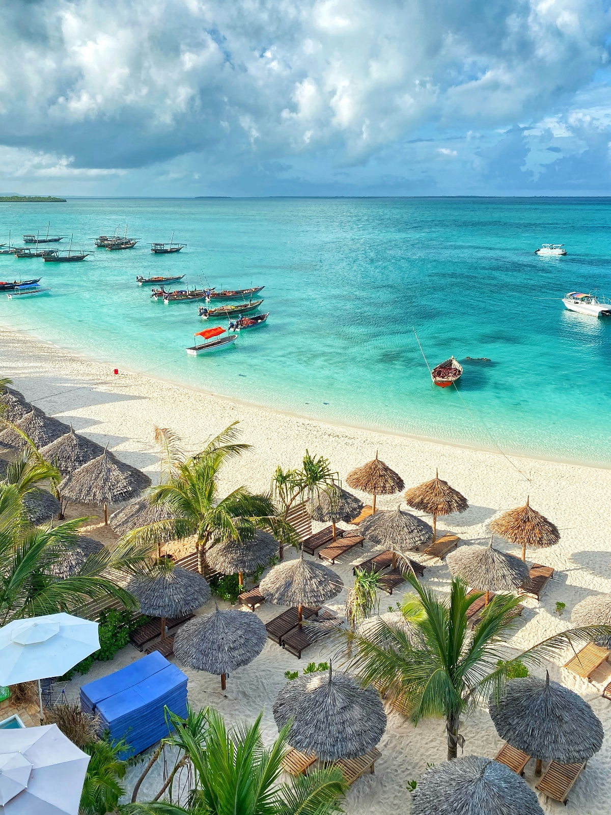 A tropical beach scene shows white sand lined with thatched palm-frond umbrellas and wooden lounge chairs, with palm trees interspersed throughout the beach area. The shallow turquoise waters contain numerous traditional wooden boats, some with canopies, anchored near the shore, while a few modern white vessels are visible farther out in the deeper blue water. The view is captured from an elevated vantage point under a cloudy sky, with a distant landmass visible on the horizon.