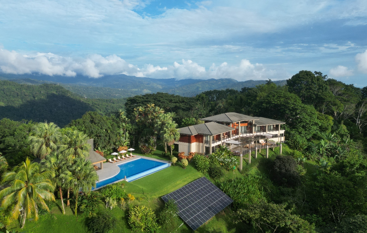 A luxury eco-lodge surrounded by lush tropical forest, featuring a modern main building with open terraces, an infinity pool with lounge chairs, and a solar panel array on a grassy hillside. Rolling green mountains stretch into the distance under a partly cloudy sky.
