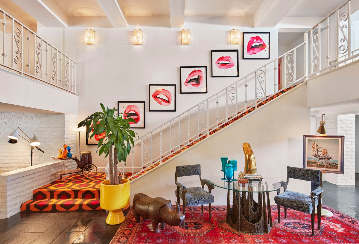 A bold, eclectic interior featuring a white staircase with ornate railings and a vibrant orange geometric patterned runner, decorated with a gallery wall of framed red lip artworks ascending along the wall. The space includes a seating area with wicker chairs around a glass table on a red patterned rug, complemented by a large plant in a yellow planter and various decorative objects creating a maximalist design aesthetic.