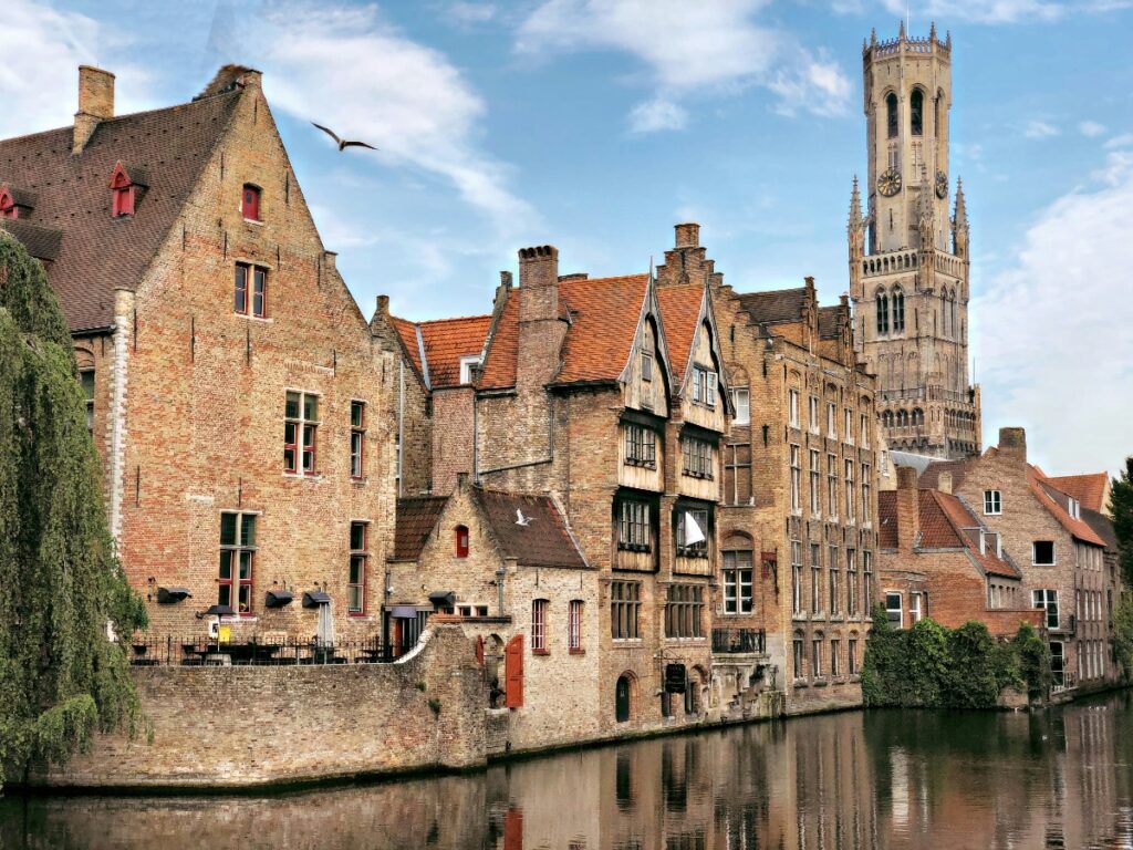 A row of historic brick buildings with stepped gables and orange-tiled roofs lines a calm canal, their facades reflected in the water below. A tall Gothic bell tower with ornate stonework rises prominently above the roofline on the right side of the scene. A bird flies through the blue sky dotted with white clouds above the medieval architecture.