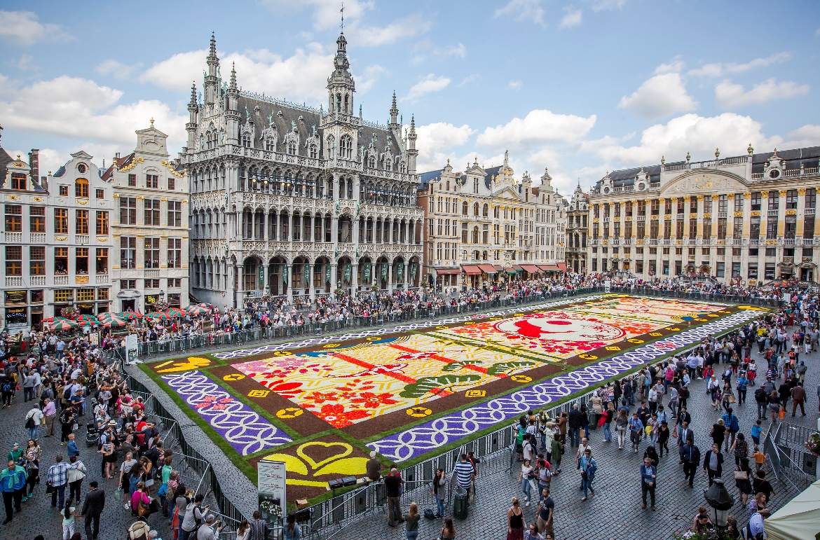 A large ornate floral carpet covers the cobblestone square of Brussels' Grand Place, featuring intricate geometric and botanical patterns in vibrant colors including red, yellow, purple, and pink. Crowds of people stand behind barriers surrounding the display, observing from all sides of the square. The historic Gothic architecture of the Maison du Roi and ornate guild houses frame the scene under a partly cloudy blue sky.