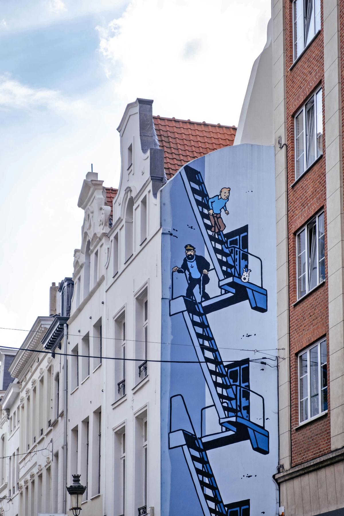 A large mural painted on a narrow building facade depicts cartoon-style figures in blue tones climbing interconnected staircases and ladders in an impossible geometric arrangement. The mural covers the side wall between a traditional white stepped-gable building on the left and a red brick building on the right. The artwork features characters and a small dog navigating the surreal architectural elements against a light blue background under a cloudy sky.