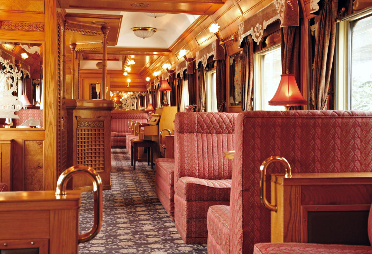 A luxury train dining car features rows of booth-style seating upholstered in quilted rose-colored fabric, arranged along both sides of a central aisle with patterned carpeting. The interior is richly appointed with ornately carved wood paneling, brass railings and fixtures, table lamps with pink shades, ceiling-mounted globe lights, and windows dressed with layered curtains that line the length of the car.