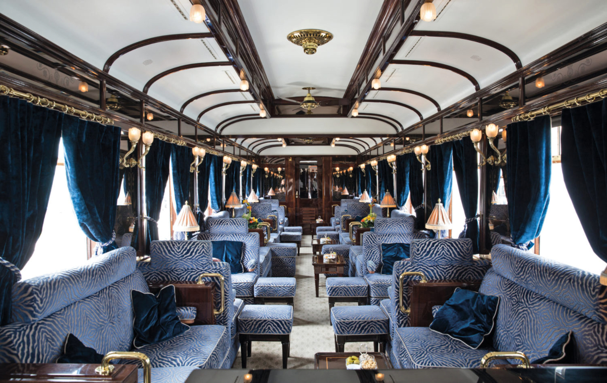 A luxury train observation car features symmetrical rows of upholstered armchairs with zebra-pattern fabric in blue and grey tones, arranged along both sides of a central aisle with matching ottomans and small side tables. The interior is decorated with dark wood paneling, heavy blue velvet curtains covering large windows, ornate brass wall-mounted light fixtures, and an elaborately detailed white arched ceiling with decorative molding and dark wood beams.