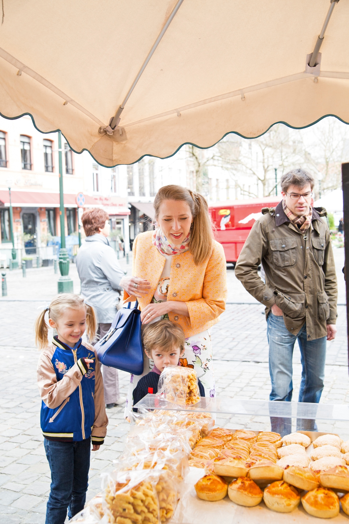 A family of four stands at an outdoor bakery stall under a beige awning, with a woman in a yellow cardigan helping two young children select from a table display of pastries, rolls, and baked goods. A man in an olive jacket and glasses stands nearby, while other pedestrians and European-style buildings are visible in the background. The scene takes place on a cobblestone plaza or street with a red vehicle visible in the distance.