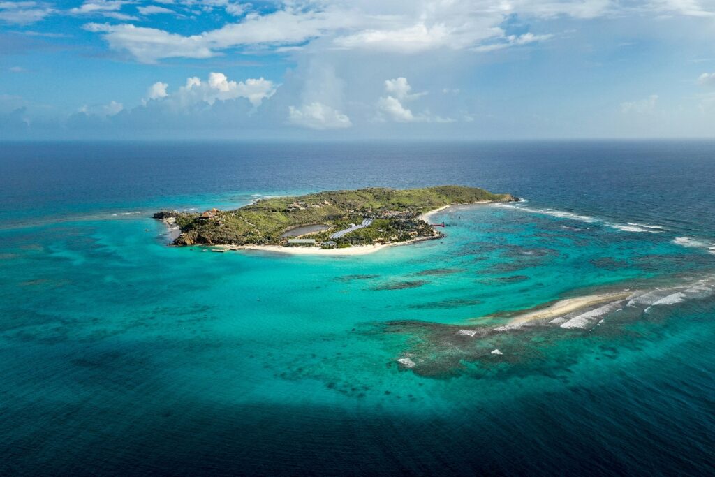 An aerial view of a small tropical island covered with green vegetation and white sandy beaches, surrounded by shallow turquoise waters with visible coral reef formations and darker patches. The island sits in a lagoon encircled by a reef where white waves break, with the water transitioning from bright aqua in the shallows to deep blue in the open ocean beyond, under a partly cloudy sky.
