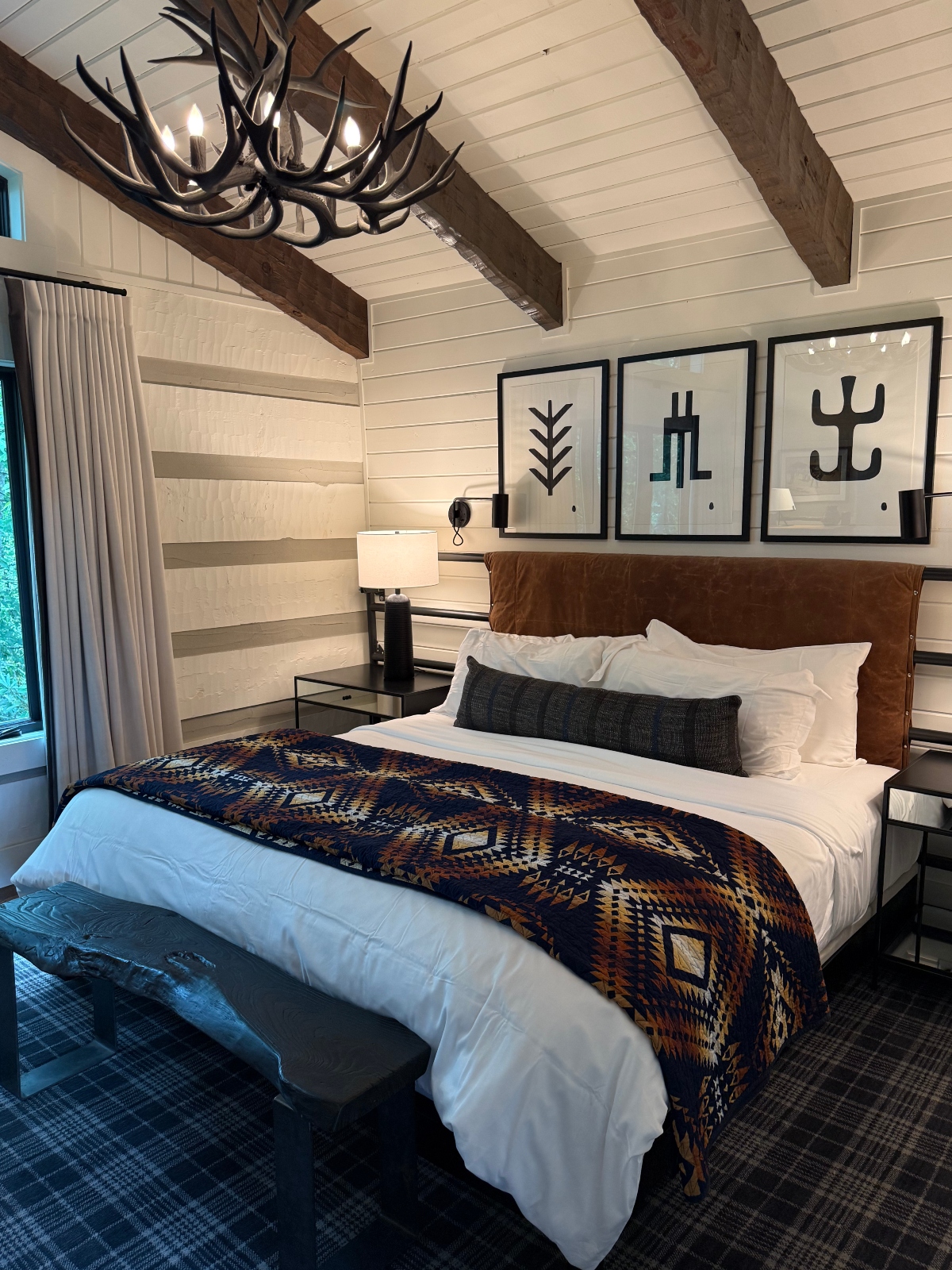 A rustic-modern bedroom features white shiplap walls with exposed wooden ceiling beams and an antler chandelier. The bed has a brown leather headboard, white linens, and a geometric-patterned throw blanket in navy, rust, and tan tones, with three black-framed artworks displaying simple geometric symbols mounted above. The room includes metal-framed nightstands with lamps, blue plaid carpeting, and a bench at the foot of the bed.