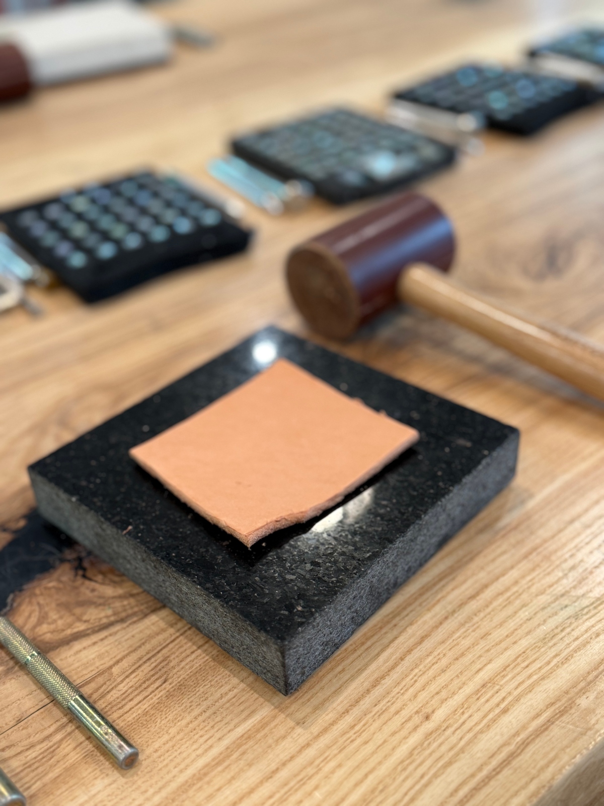 A leather crafting workspace displays a square piece of tan leather centered on a black foam pad, with a wooden mallet positioned nearby. Several leather stamping tools with metal letter or pattern stamps are visible in the background, along with a metal stylus in the foreground. The tools are arranged on a wooden work surface, appearing ready for leather stamping or tooling work.