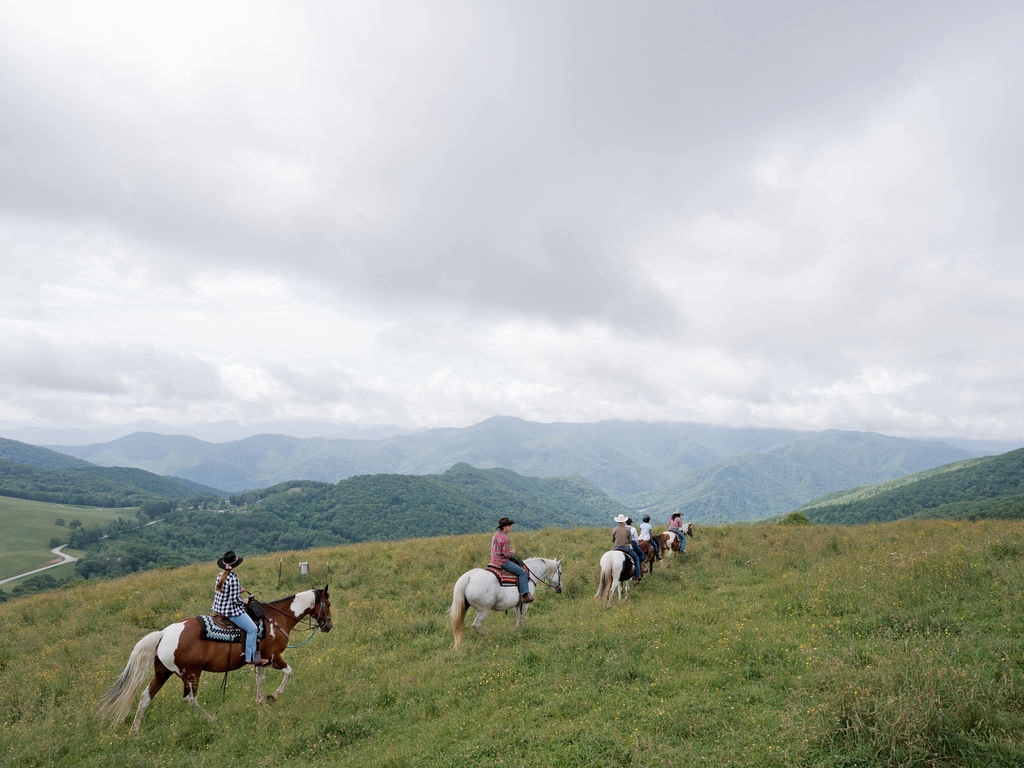 A line of horseback riders traverses a grassy mountain hillside, with riders mounted on horses of varying colors including brown and white. The trail crosses an open meadow with layers of forested mountains visible in the distance under an overcast sky. The scenic mountain landscape features multiple ridges receding into the background.