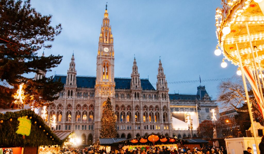 A Gothic Revival building with a prominent central spire and clock tower stands illuminated against the evening sky, with four smaller matching spires flanking the main structure. In front of the building, a large Christmas market bustles with crowds of people among wooden market stalls, featuring a tall decorated Christmas tree and carnival rides including a brightly lit carousel on the right side.