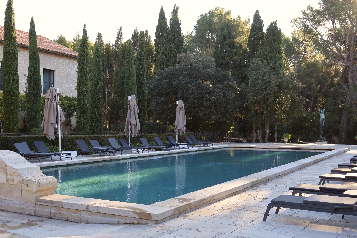A rectangular swimming pool with stone coping is surrounded by dark lounge chairs and furled beige umbrellas on a paved stone deck. Tall cypress trees and mixed vegetation create a backdrop behind the pool area, with a stone building featuring a terracotta roof visible on the left side. A classical statue can be seen in the distance among the trees on the right.