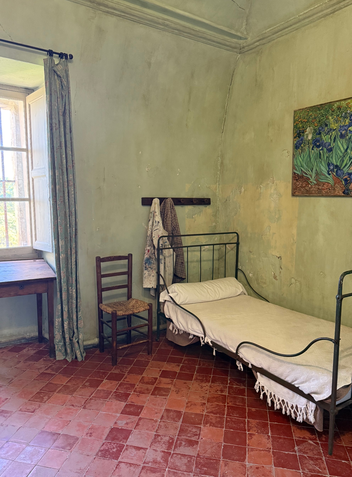 A sparse bedroom features a simple metal-framed bed with white linens, a wooden chair with a woven seat, and weathered pale green walls with visible aging and texture. Terra cotta tile flooring extends throughout the space, and an open window with blue-gray curtains allows natural light to enter. A framed print of irises hangs on the wall near the bed, and garments are draped on wall-mounted hooks.