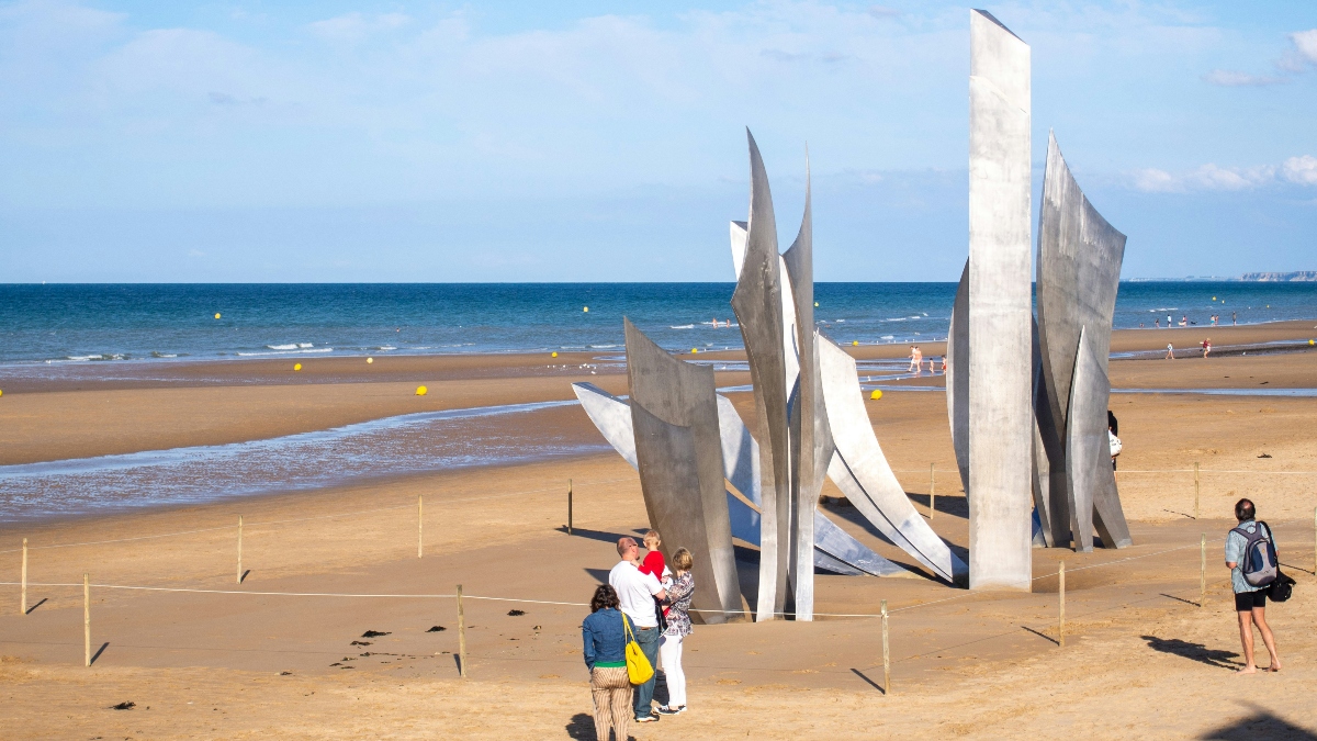 A family and a man with a backpack observe a large metal sculpture on a sandy beach. The sculpture consists of several tall, curved, silver-colored abstract shapes arranged in a circle. The beach extends into the background with calm ocean waves under a partly cloudy blue sky.