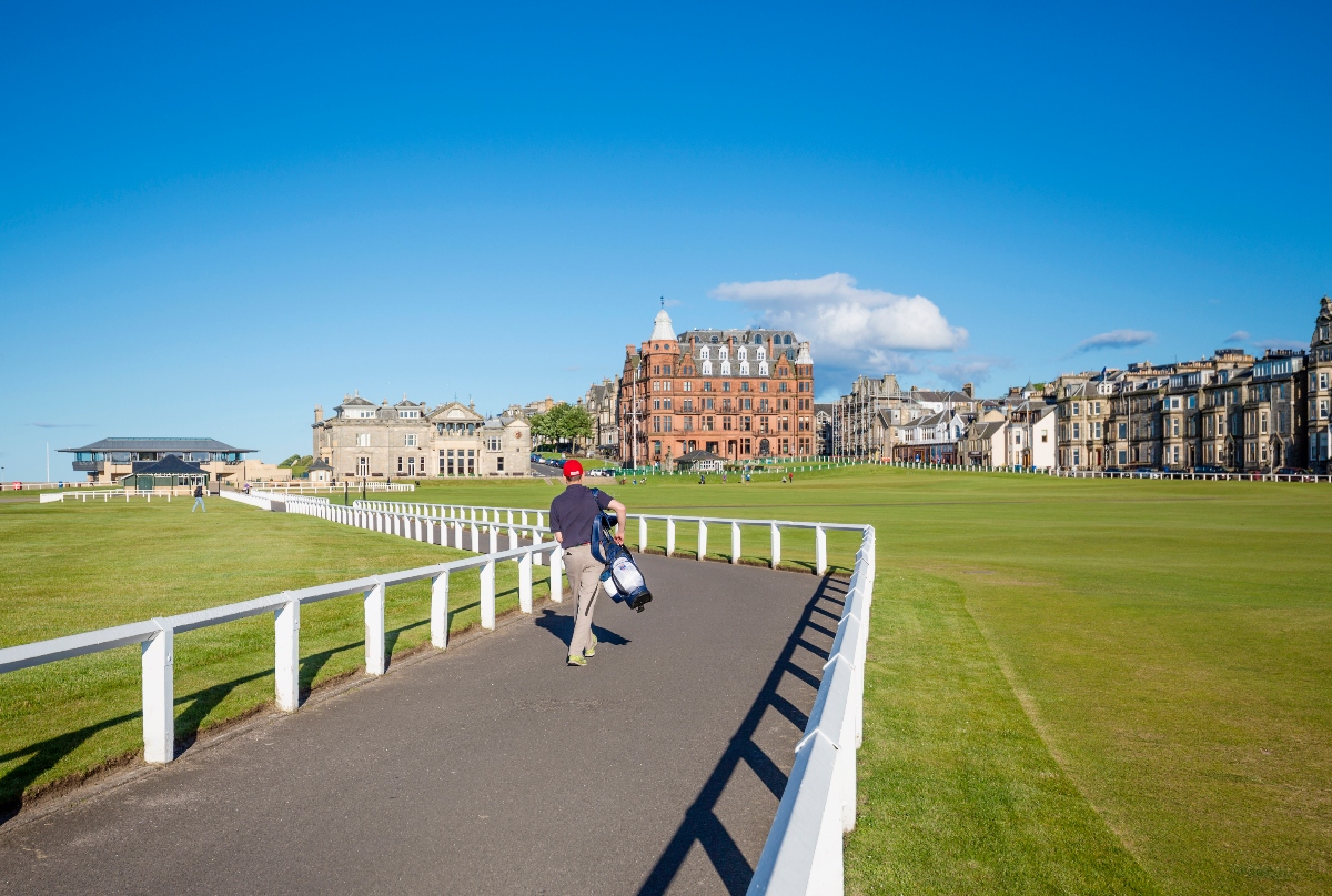 A person carrying a golf bag walks along a paved pathway bordered by white railings, with expansive green lawns on either side. In the background, a row of historic multi-story buildings with varied architectural styles lines the horizon under a clear blue sky with scattered white clouds.