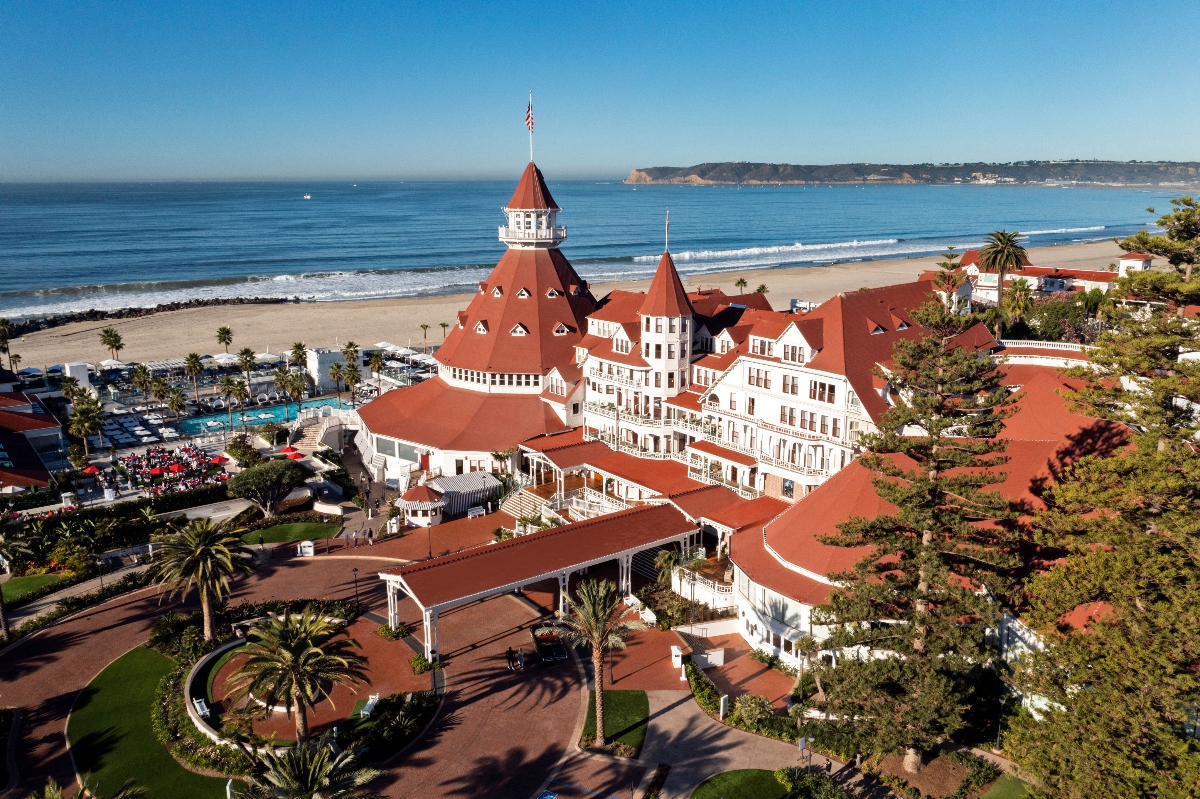 An aerial view shows a large Victorian-style resort hotel with distinctive red tile roofs and white walls, featuring a prominent conical tower topped with a cupola and American flag. The grand multi-story building sits directly on a sandy beach with the Pacific Ocean beyond, surrounded by palm trees and manicured grounds that include what appears to be a swimming pool area. The coastline curves along the horizon under a clear blue sky, with distant hills visible across the water.