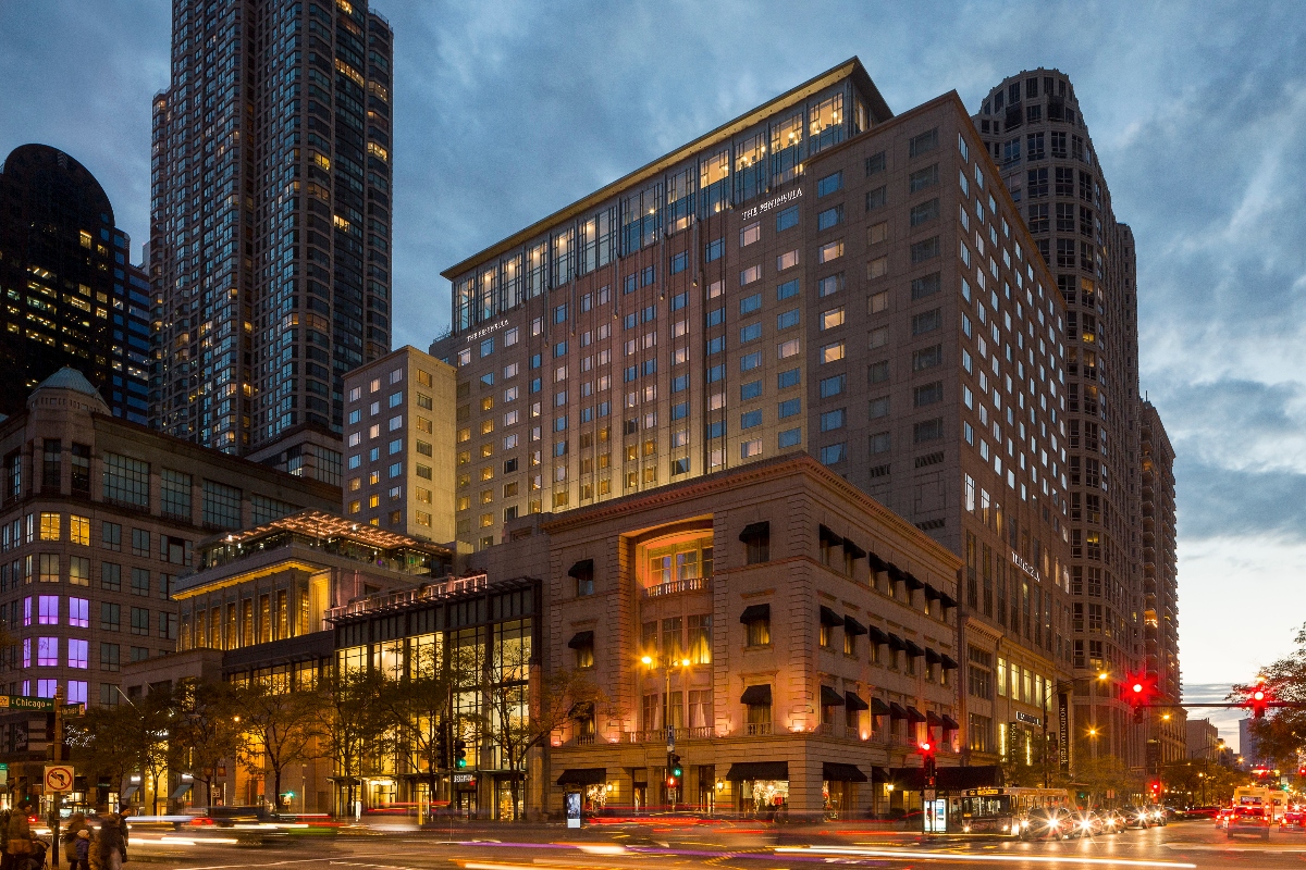 A cityscape photograph taken during dusk shows a mix of modern high-rise buildings and classical architecture along a busy urban street. The scene captures illuminated building windows against a blue twilight sky, with light trails from moving vehicles creating streaks on the street below. Traffic lights, street lamps, and the warm glow from building interiors create a vibrant evening atmosphere in this metropolitan setting.