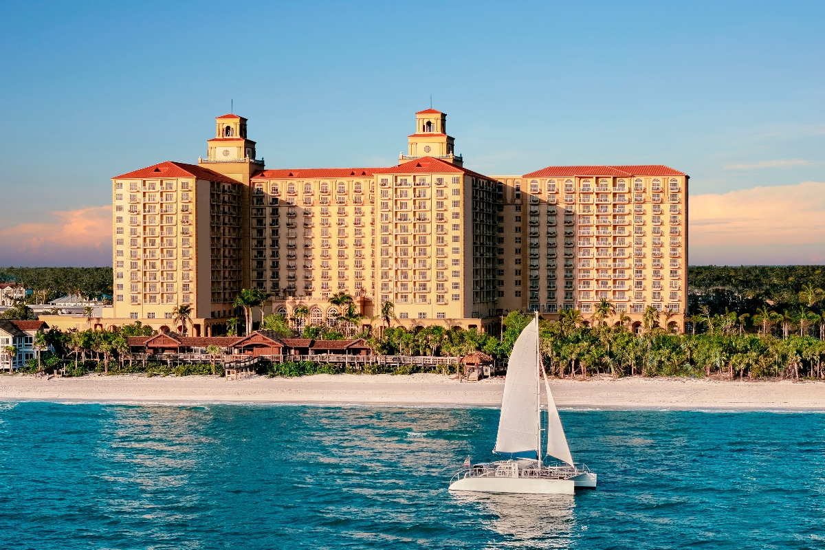 A large beachfront resort hotel with distinctive twin towers topped by red tile roofs sits on a white sandy beach surrounded by palm trees and tropical vegetation. In the foreground, a white catamaran sailboat floats in clear turquoise waters under a blue sky. The multi-story building features Mediterranean or Spanish colonial-style architecture and extends along the coastline.