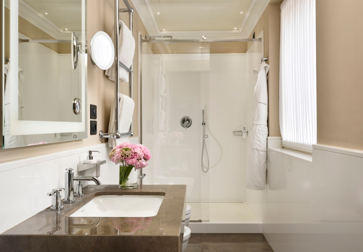 A luxury bathroom features a marble vanity with an undermount sink, large illuminated mirror with an attached magnifying mirror, and a glass-enclosed shower with marble walls. The space includes white bathrobes hanging on hooks, pink flowers in a vase on the counter, and neutral beige tones throughout with recessed ceiling lighting.