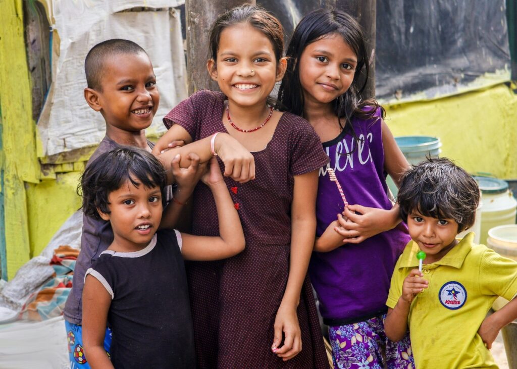 This image shows a group of five children of varying ages posing together and smiling at the camera. The children are wearing different colored clothing including purple, yellow, and dark-colored shirts, with one child wearing what appears to be a polka-dotted dress. They are positioned in front of a background that includes yellow walls and various containers, suggesting an outdoor residential or community setting.
