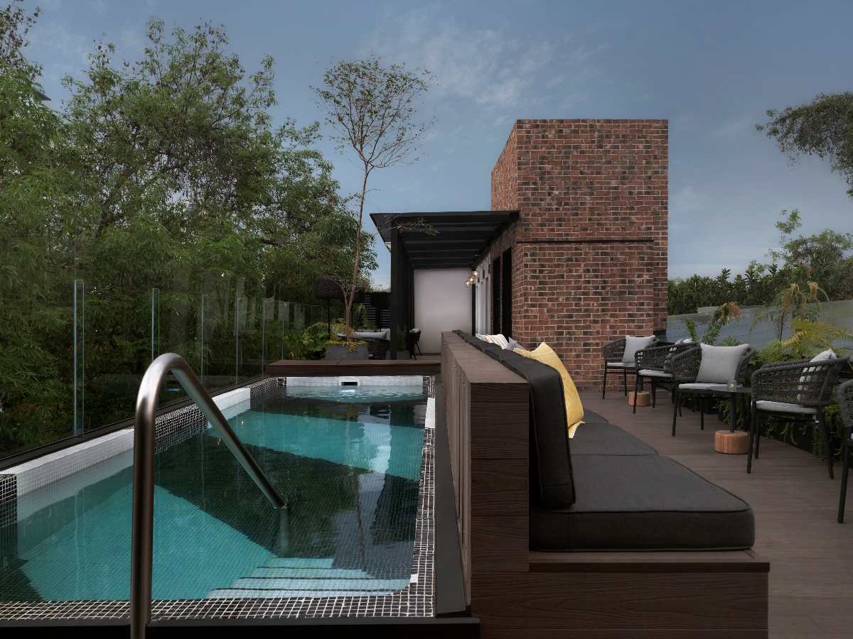 A rooftop terrace featuring a rectangular swimming pool with turquoise water, white mosaic tile trim, and a metal ladder, surrounded by dark wood decking. Adjacent to the pool is a modern outdoor lounge area with a daybed topped with black cushions and a yellow throw, wicker seating with gray cushions, and a prominent red brick chimney structure. The space is enclosed by glass railings with mature trees visible beyond, creating a private urban retreat under an overcast sky.