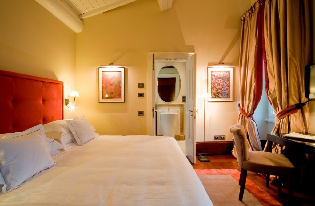 This image shows an elegant hotel room or bedroom featuring a bed with white linens and a red upholstered tufted headboard, set against warm yellow walls with exposed white ceiling beams. The room includes wall-mounted reading lamps, two framed artworks, and a bathroom door with an oval mirror visible in the center. Heavy gold and burgundy curtains frame a window on the right side, while a wooden chair sits in the foreground on dark hardwood flooring.