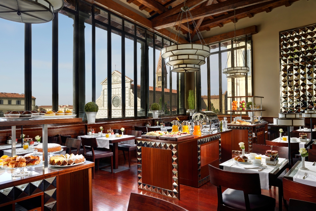 This image shows an upscale restaurant dining room with floor-to-ceiling windows offering views of historic buildings including a prominent church or cathedral. The space features exposed wooden ceiling beams, modern tiered pendant lighting, and a breakfast buffet setup with pastries and beverages displayed on wooden stations with geometric patterned trim. Dark wood furnishings, white-linened tables, and polished floors complete the elegant interior that blends traditional and contemporary design elements.