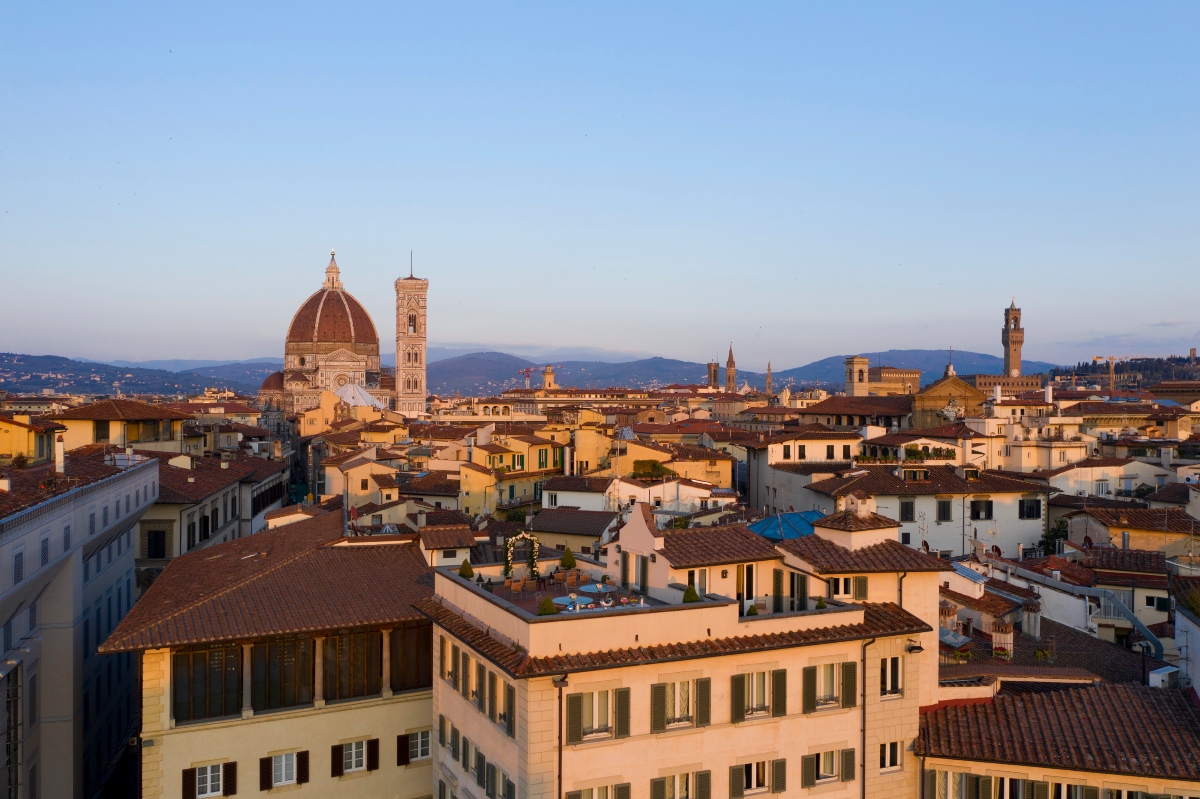 This aerial view shows a historic Italian city with traditional terracotta-roofed buildings spread across the urban landscape, dominated by a prominent cathedral featuring a large Renaissance dome and tall bell tower. The cityscape extends toward rolling hills visible in the distance under a clear blue sky, with the warm lighting suggesting either sunrise or sunset. The architecture displays a mix of residential buildings, historic structures, and notable landmarks characteristic of a medieval Italian city center.