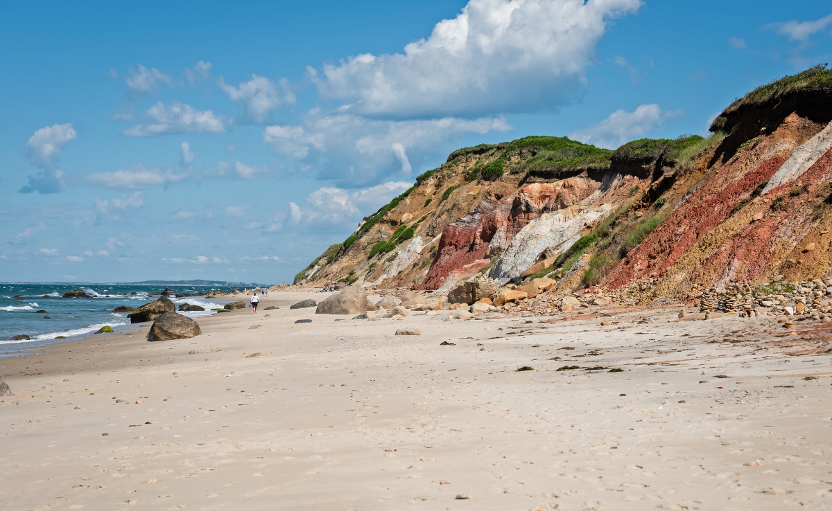 A sandy beach stretches along the shoreline with ocean waves visible on the left and two small figures walking in the distance. Dramatic multicolored coastal cliffs rise on the right side, displaying layers of red, white, and brown sedimentary rock topped with green vegetation, with scattered boulders at their base.