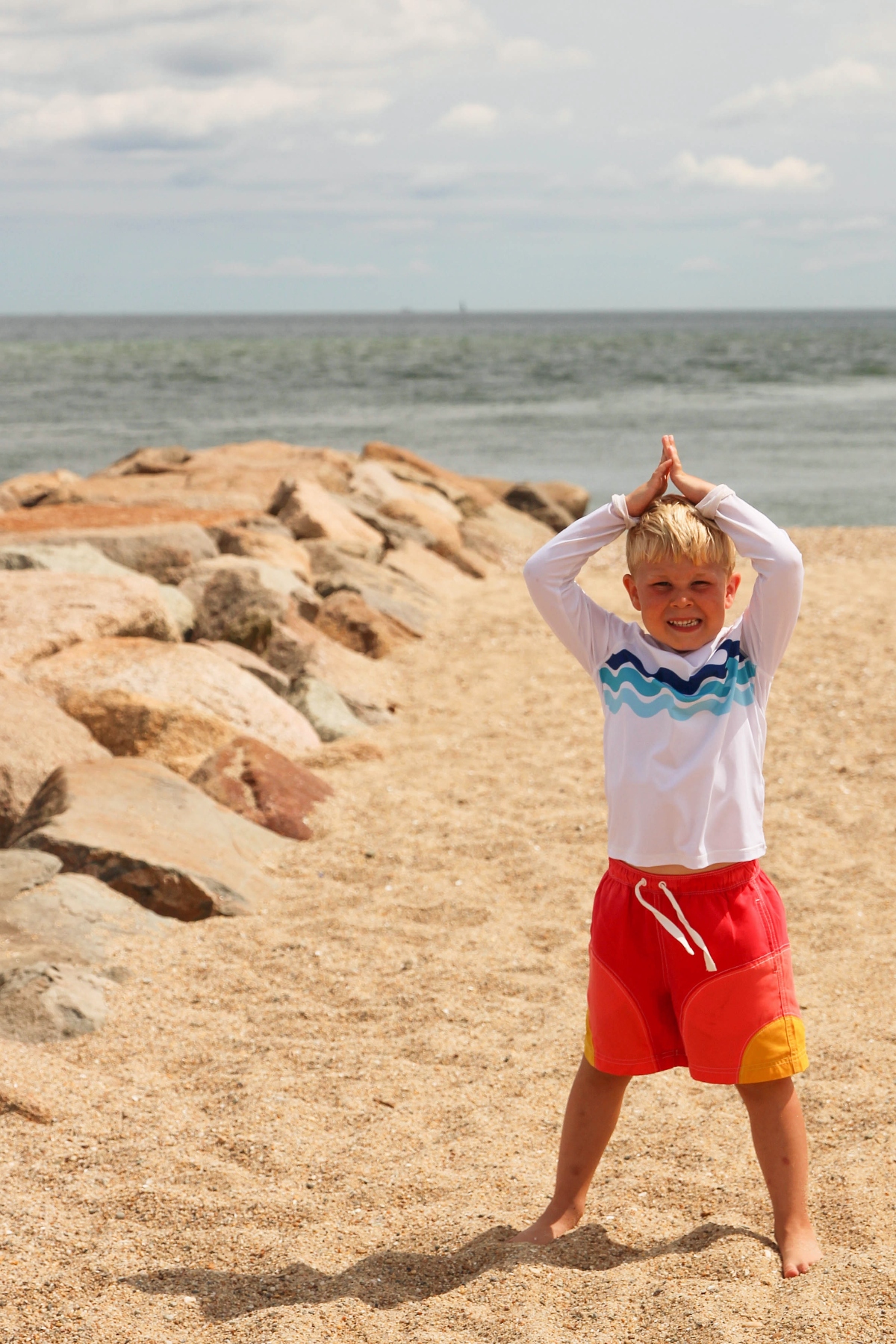 A young child wearing a white long-sleeved shirt and red shorts stands on sandy ground with both arms raised above their head and palms pressed together. The scene is set on a beach with large rocks or boulders in the middle ground and an ocean horizon under a cloudy sky in the background.
