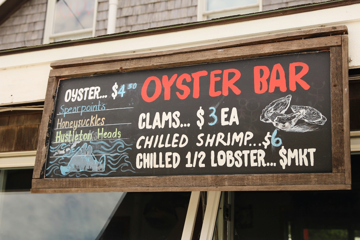 A wooden-framed chalkboard menu sign displays "OYSTER BAR" in large red letters at the top, listing seafood items and prices including oysters ($4.50), clams ($3 EA), chilled shrimp ($6), and chilled half lobster (market price). The black menu board features white chalk illustrations of oyster shells and blue wave designs, and is positioned in front of a building with shingled siding.