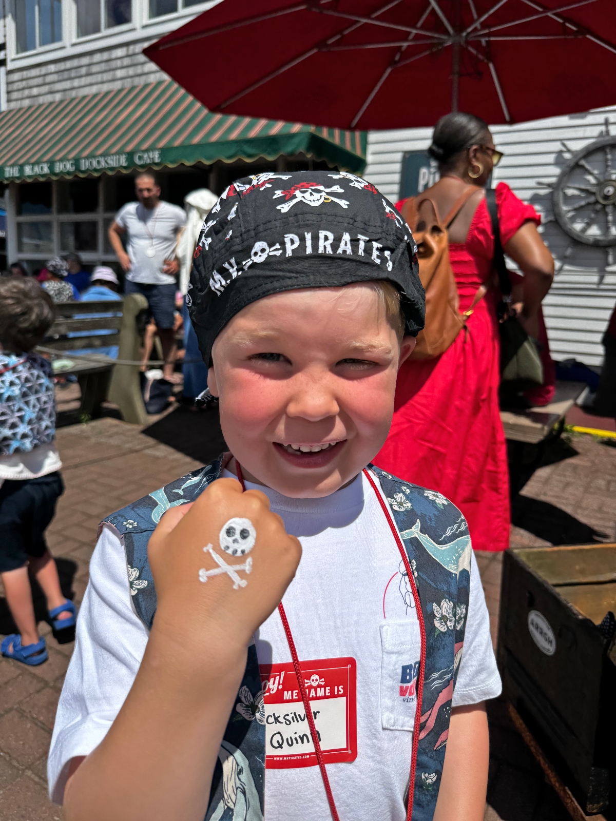 A young child wearing a black pirate bandana with skull and crossbones design smiles at the camera while displaying a small skull and crossbones temporary tattoo on their raised fist. The child is dressed in pirate-themed clothing with a red name tag and stands in what appears to be an outdoor boardwalk or waterfront area with buildings, awnings, and other people visible in the background.