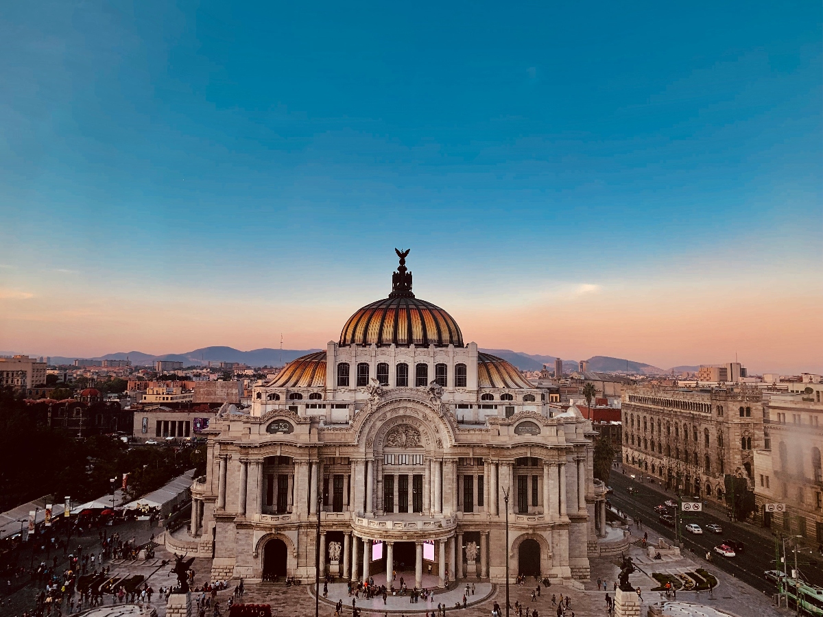 The image shows the Palacio de Bellas Artes in Mexico City at sunset, with its distinctive domed roof featuring orange and yellow tiles. The neoclassical and art nouveau building is surrounded by city streets and people walking in the plaza. The background includes distant mountains under a clear blue-to-pink gradient sky.