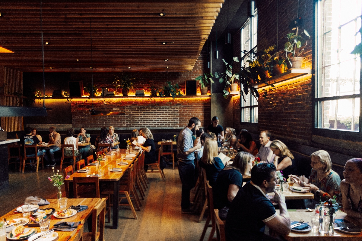 A modern restaurant interior features multiple wooden communal tables with diners seated throughout the space, illuminated by warm lighting beneath a slatted wood ceiling. The dining room has exposed brick walls, large windows on the right side, and a backlit shelf displaying potted plants along the rear wall.