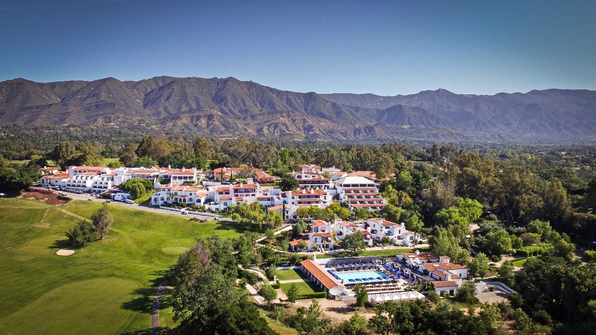 An aerial view shows a sprawling resort complex with white Mediterranean-style buildings featuring red tile roofs, scattered across rolling hills and connected by winding pathways. The property includes a prominent golf course with manicured green fairways in the foreground, along with what appears to be a central pool and recreation area surrounded by guest accommodations. The entire resort is set against a dramatic backdrop of rugged mountains under a clear blue sky, with dense vegetation and mature trees interspersed throughout the grounds.