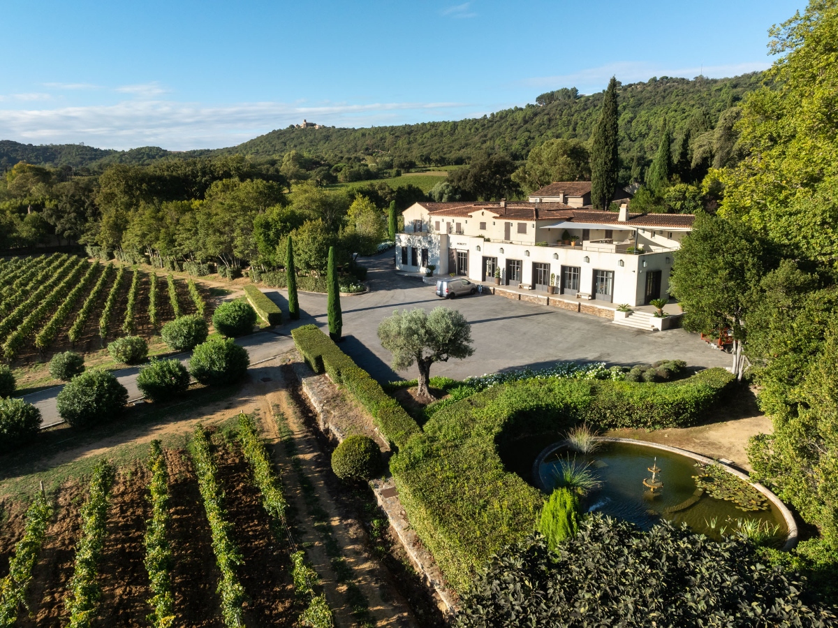 An aerial view shows a large Mediterranean-style building with terracotta roofing situated on a hillside property surrounded by dense green forests and rolling hills. The estate features organized vineyard rows on the left side, a circular pond with a fountain in the foreground, cypress trees, landscaped gardens, and a paved courtyard area, all set against a blue sky with scattered white clouds.