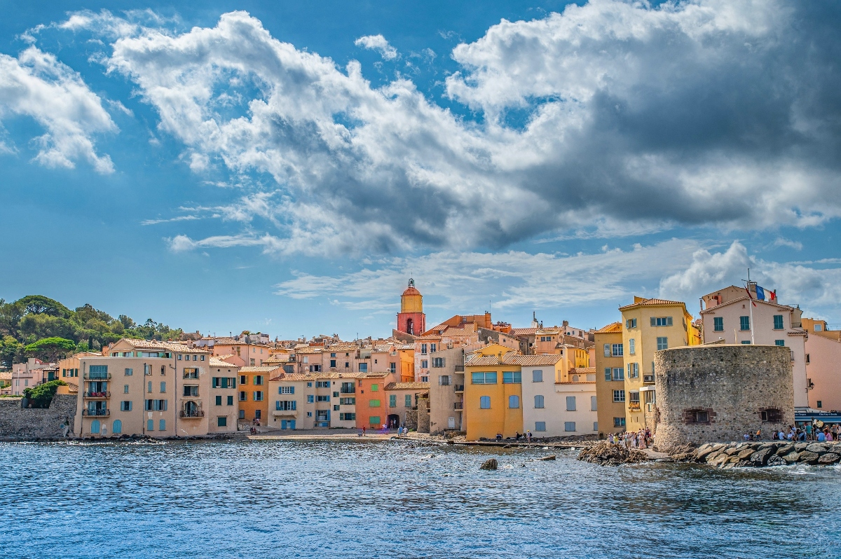 A Mediterranean coastal town displays rows of colorful multi-story buildings in shades of yellow, orange, pink, and cream along the waterfront, with a prominent bell tower rising above the rooflines. The scene is viewed across calm blue water under a dramatic sky filled with white and gray clouds, with a stone fortification visible on the right side and wooded hills in the background.