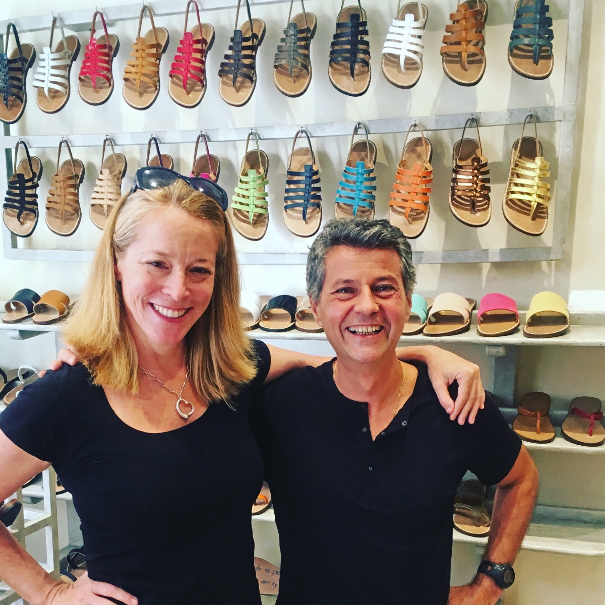 Two people pose together with their arms around each other in a shoe store, both wearing black tops and smiling at the camera. Behind them are multiple white shelves displaying rows of colorful sandals in various styles and bright colors including pink, blue, orange, white, and black.