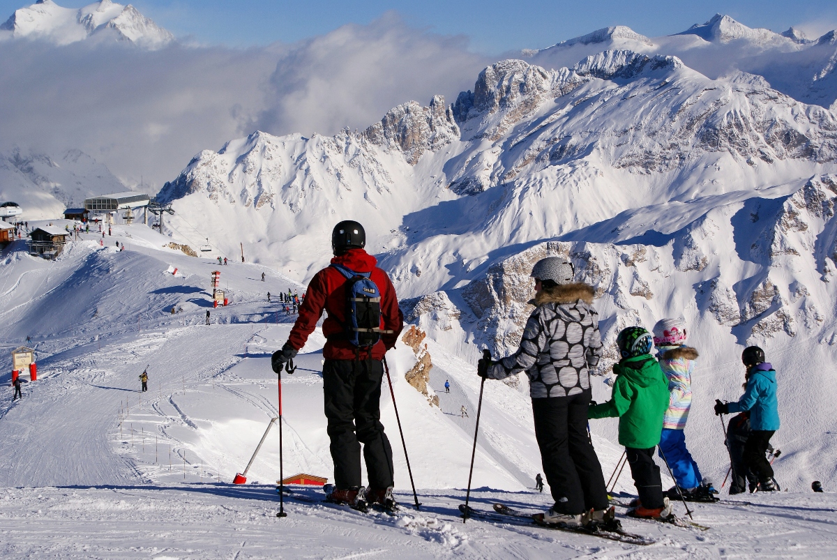 A group of skiers, including an adult in a red jacket with a backpack and several children in various colored jackets, stand at the top of a ski slope looking out at dramatic snow-covered Alpine peaks. In the background, jagged mountain summits rise against a blue sky with wispy clouds, while ski lifts and a mountain lodge are visible on the left side of the slope. Other skiers can be seen descending the groomed trails below.