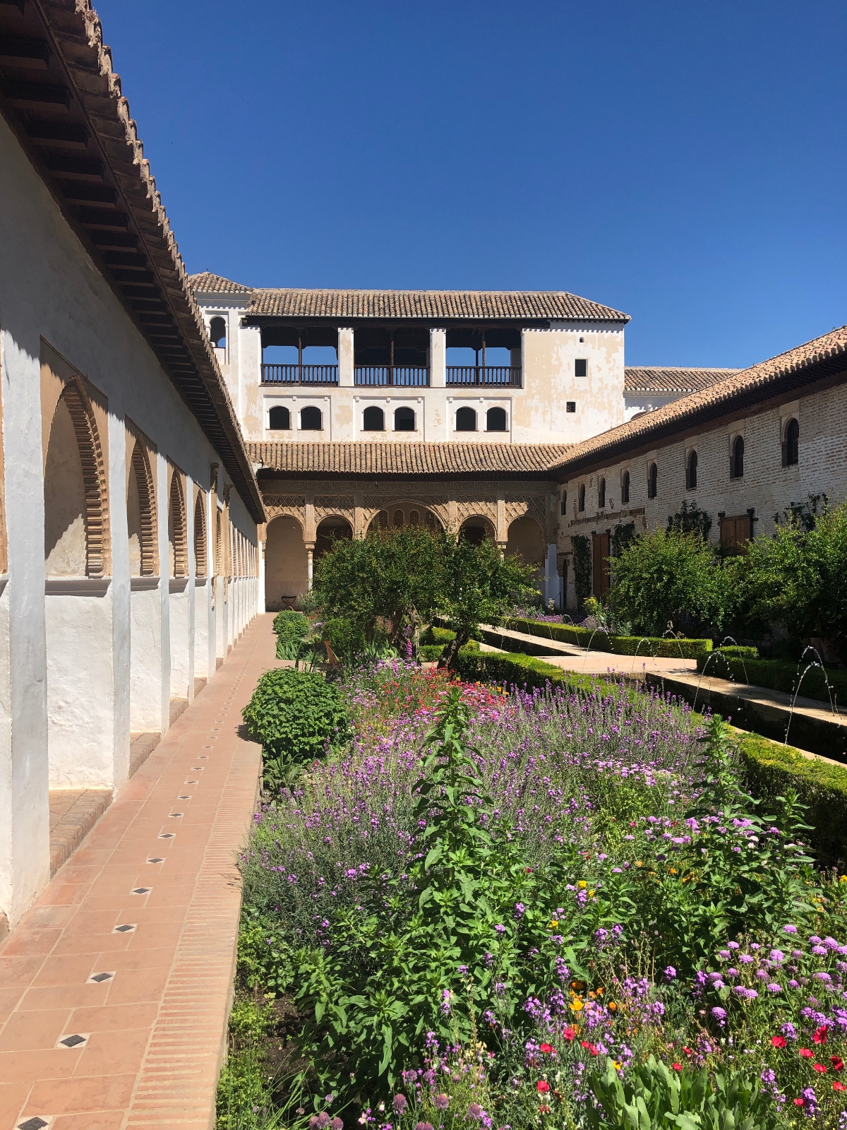 A historic courtyard features Moorish-style architecture with white arched colonnades, terracotta tile roofs, and a multi-level building with wooden balconies in the background. The central garden area contains formal landscaping with colorful flowering plants in purple, pink, and red, geometric hedge patterns, and a series of water fountains along a rectangular pool.