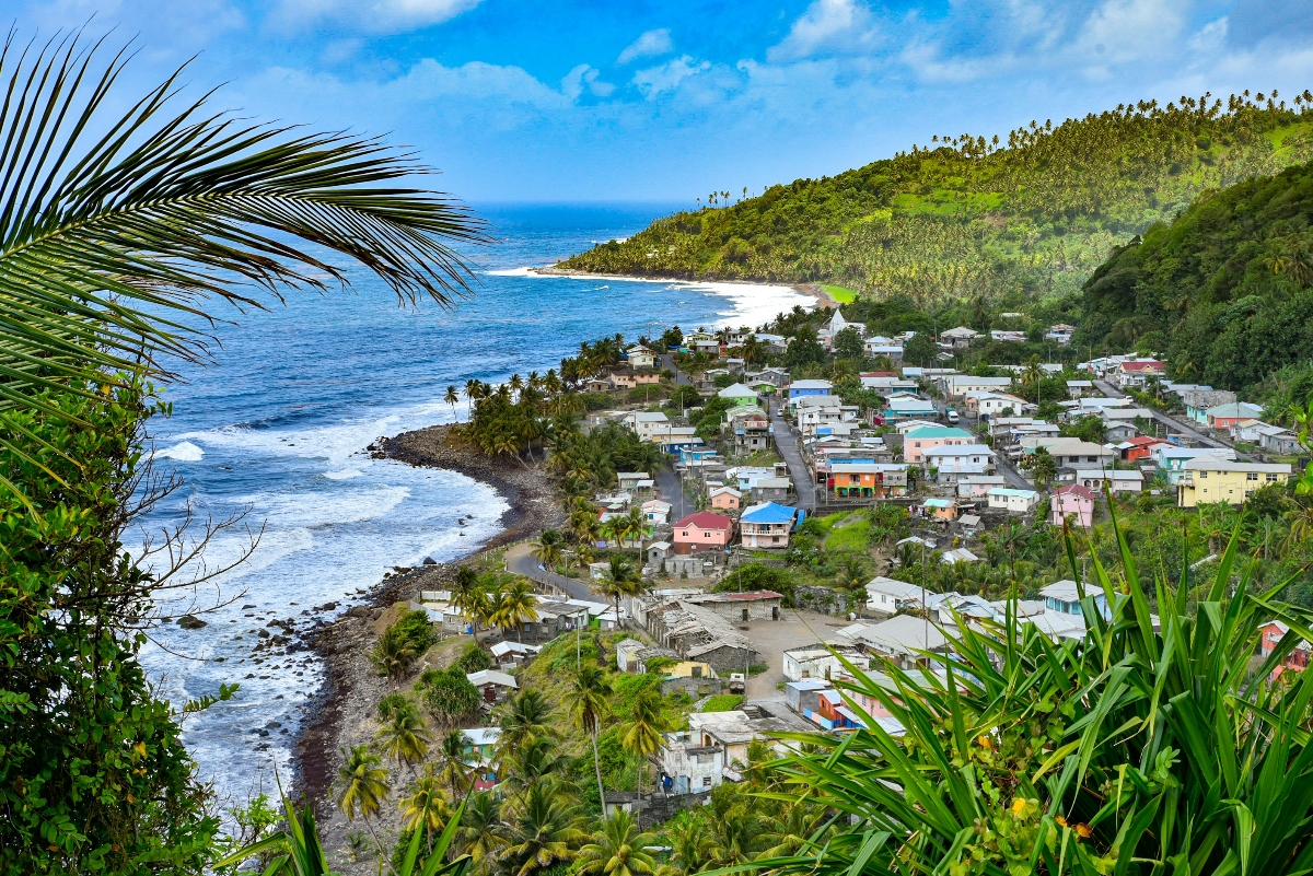 A coastal village with colorful houses nestled between lush, tropical green hillsides and the ocean, with palm trees dotting the landscape and palm fronds framing the foreground. The settlement sits along a dark rocky shoreline where blue ocean waters meet the land, with waves visible along the coast and partly cloudy skies above.
