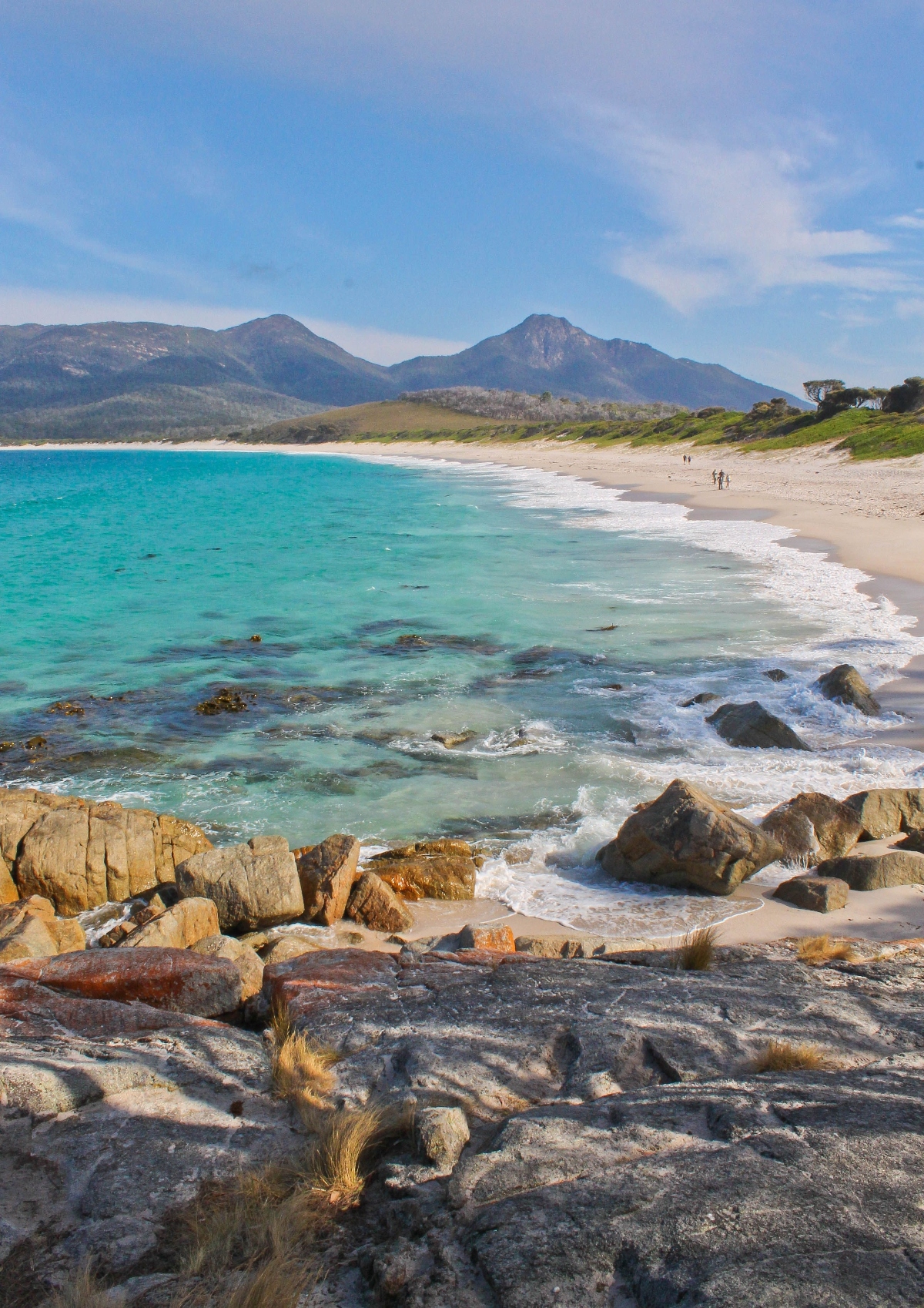A pristine white sand beach curves along turquoise waters, with scattered rocks visible both in the clear water and along the shoreline. The view is captured from an elevated rocky vantage point, showing a few small figures of people walking on the expansive beach. In the background, mountain ranges rise behind green coastal vegetation under a partly cloudy blue sky.