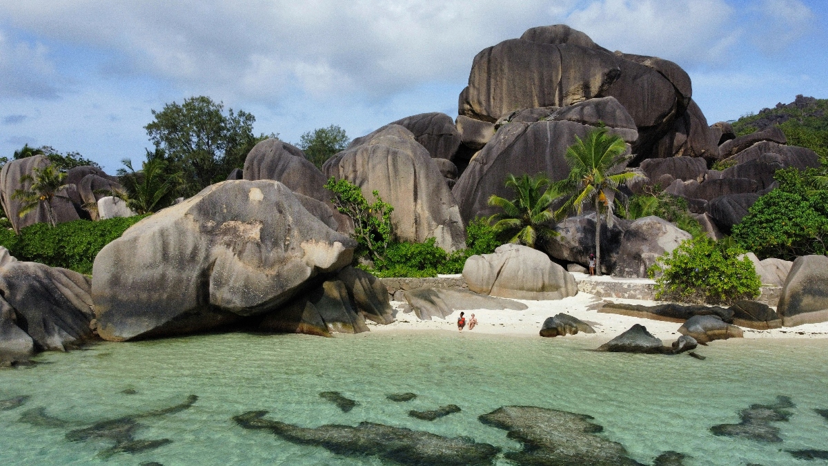 Massive, smooth granite boulders with distinctive rounded formations create a dramatic landscape around a small white sand beach with shallow turquoise water. Two people are visible on the beach, while tropical vegetation including palm trees grows among the rocks beneath a partly cloudy sky.