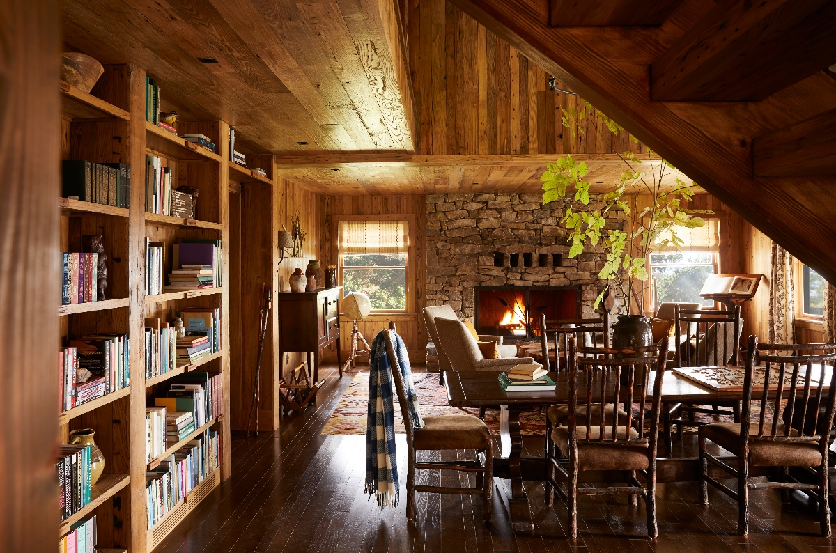 This image shows the interior of a rustic wooden room featuring floor-to-ceiling built-in bookshelves filled with books along the left wall. The space includes a stone fireplace with an active fire in the center background, a wooden dining table with chairs in the foreground, and exposed wooden beam construction throughout the ceiling and walls. Natural light enters through windows on the right side, and a leafy plant is visible near the fireplace area.