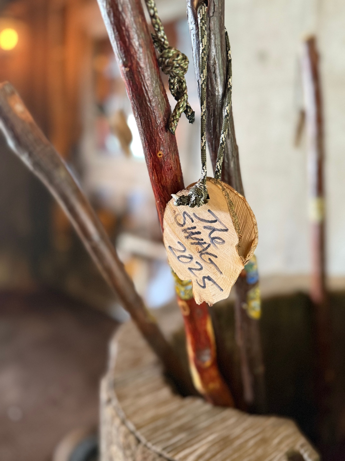 This image shows a handwritten paper tag tied with green rope or cord to one of a group of wooden hiking sticks in a barrel. The tag appears to have text written on it in what looks like pen or marker, and the background is softly blurred showing an indoor space with warm lighting. The wooden structure appears weathered or rustic in appearance.