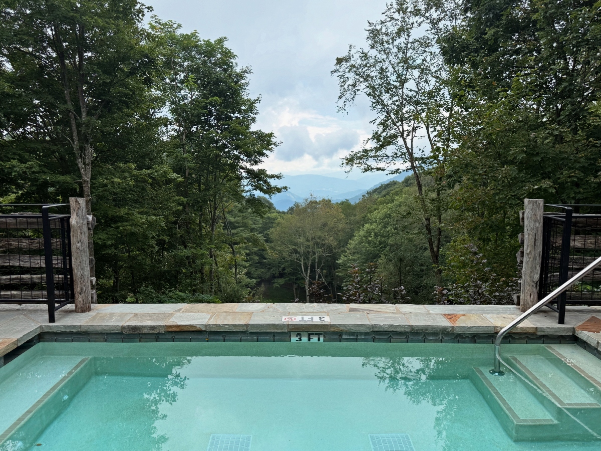 This image shows a rectangular swimming pool with clear blue-green water, featuring depth markings and a handrail, positioned on what appears to be a stone or concrete deck. The pool is surrounded by metal railings and overlooks a densely forested landscape with rolling hills or mountains visible in the background under an overcast sky. The setting appears to be elevated, providing a panoramic view of the surrounding natural terrain.