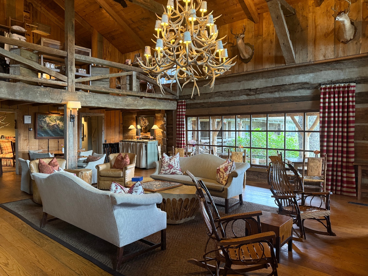 This image shows the interior of a rustic lodge or cabin featuring extensive wooden construction with exposed ceiling beams and a large antler chandelier as the centerpiece. The open-plan space includes a seating area with upholstered furniture and wooden rocking chairs, a kitchen visible in the background, and a loft level with wooden railings above. The space is decorated with mounted deer heads on the walls, red and white checkered curtains at large windows, and has hardwood flooring throughout.